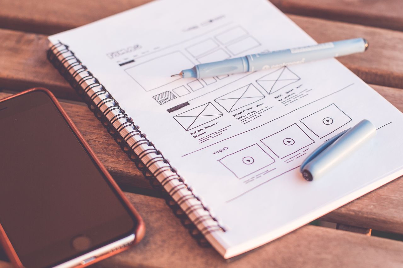 services-02 Vue rapprochée d'un carnet avec des croquis de wireframe et un smartphone posé sur un bureau en bois.