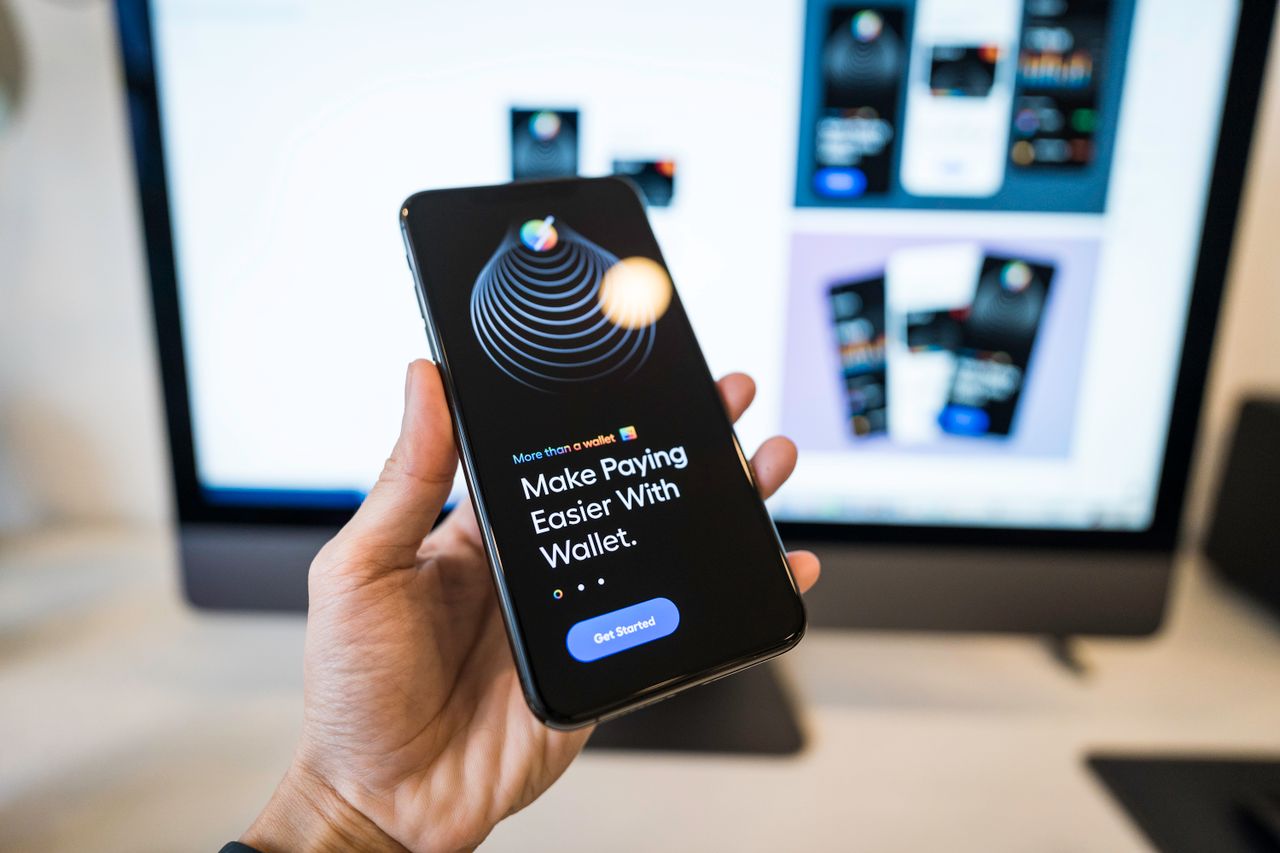 Hand holding smartphone showing a digital wallet app screen, with a blurred monitor in the background.